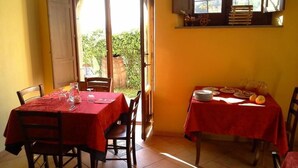 Un coin repas avec deux tables, chacune recouverte d’une nappe rouge. La pièce a des murs jaunes et une grande fenêtre avec des volets en bois.