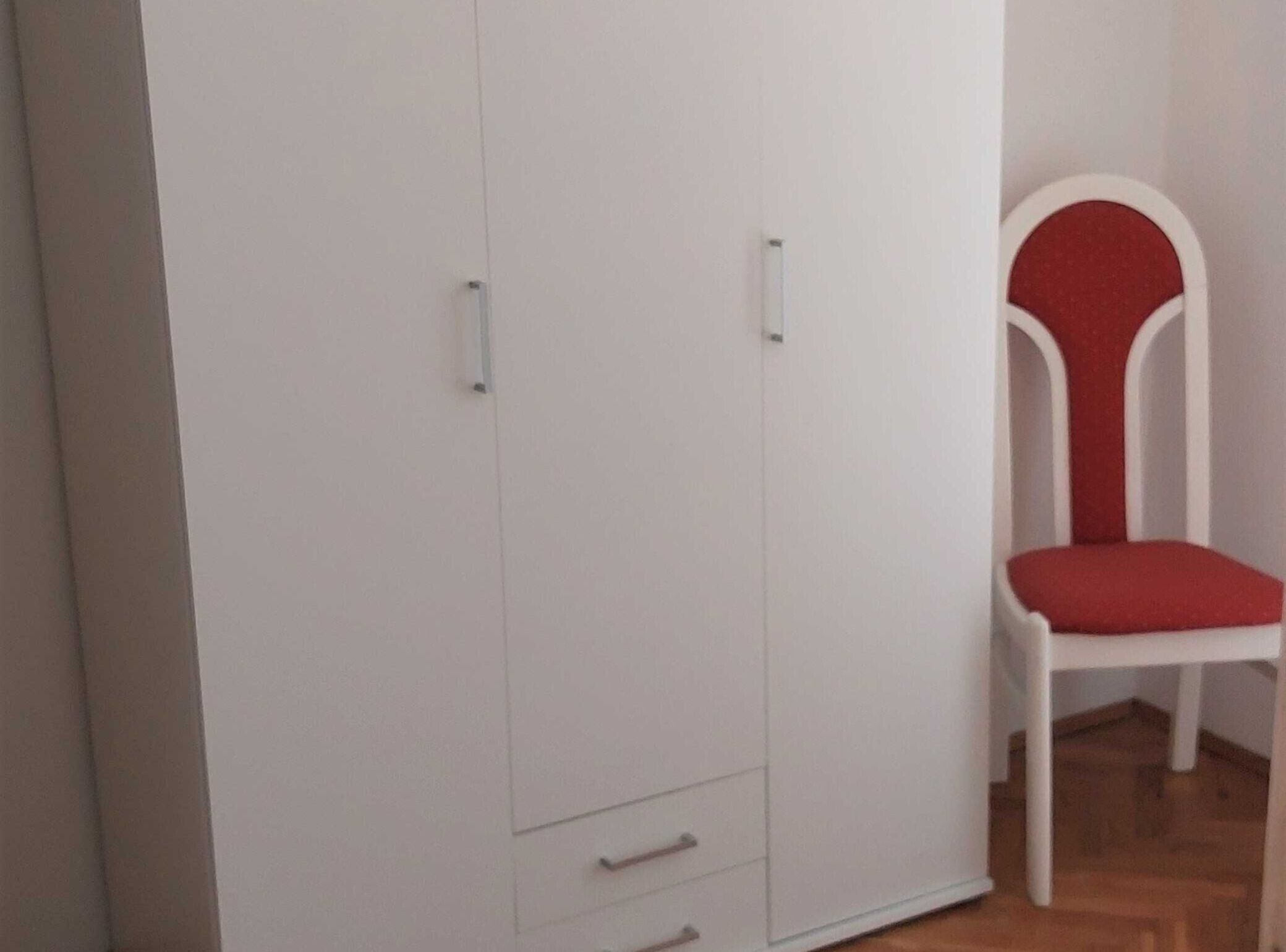 Une armoire blanche à deux portes et dotée d’un petit tiroir, une chaise rouge rembourrée et un plancher en bois.