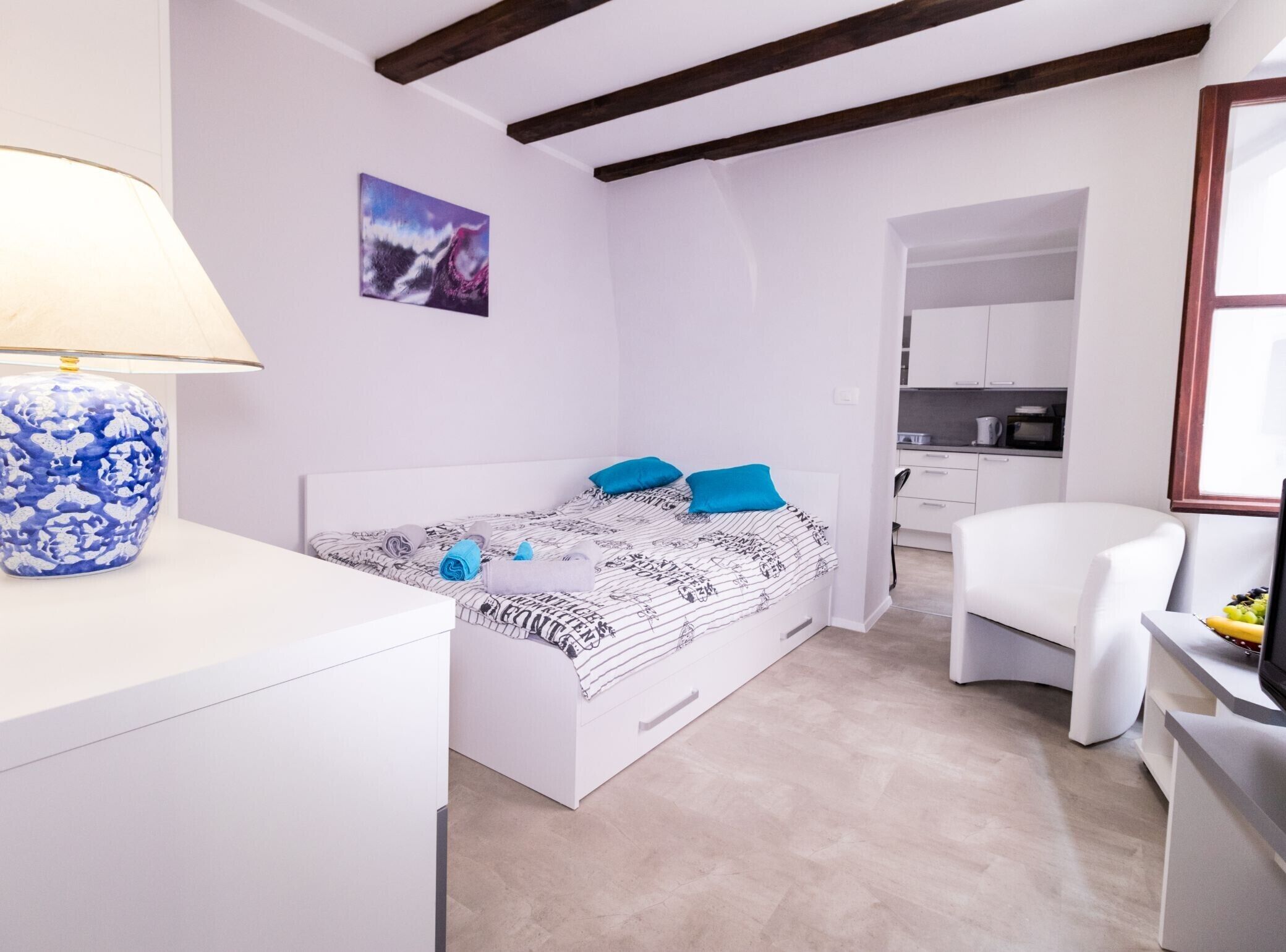 A bedroom with a bed, a chair, a desk, a lamp, and a kitchen area in the background.