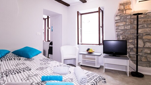 Une chambre à coucher avec un lit, une télévision, une chaise, une petite table sur laquelle se trouve un panier de fruits, et un mur en pierre.