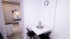 Une petite pièce blanche, avec une table blanche, deux chaises noires, une horloge au mur et une petite lampe sur une table de chevet.
