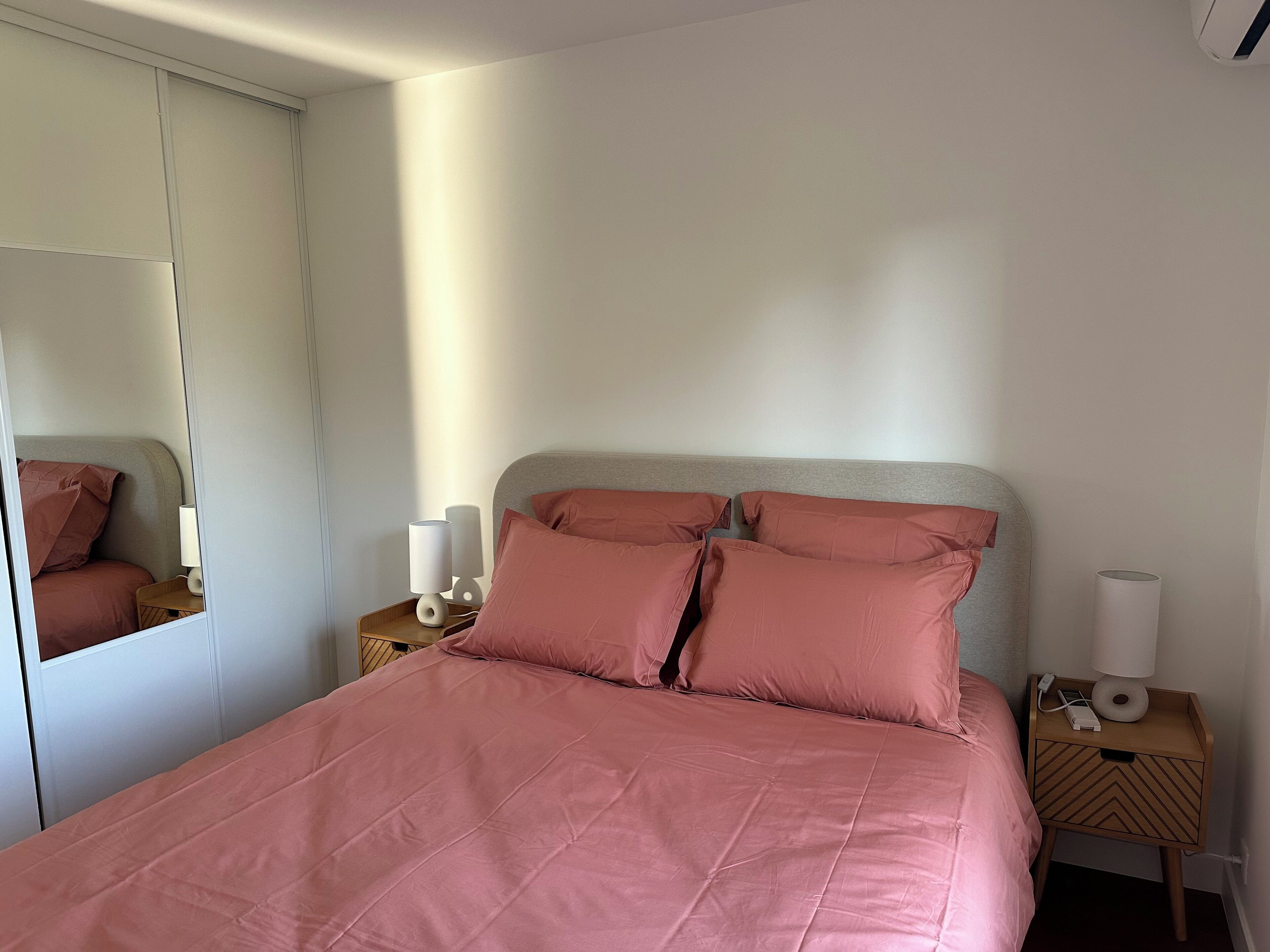 A neatly made bed with pink bedding, a bedside table with a lamp, and a mirror on the wall.