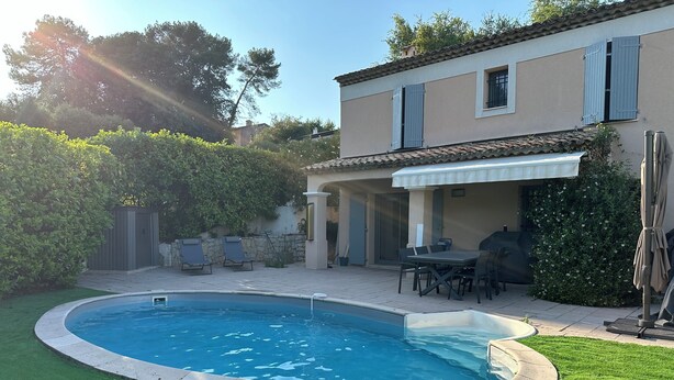 Una piscina con una zona verde circundante, un patio con mobiliario exterior y un edificio con techo de tejas.
