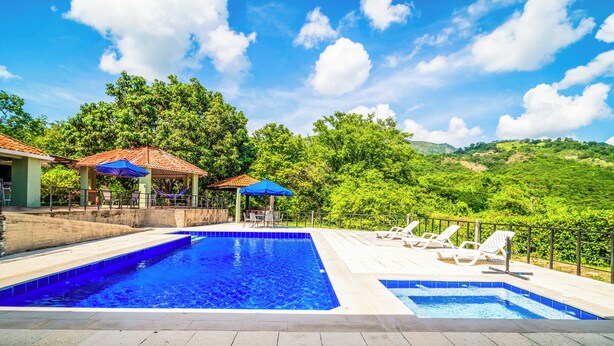 Una piscina con sillas de descanso y sombrillas, rodeada de exuberante vegetación y colinas.
