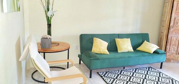 Un canapé vert agrémenté de trois coussins jaunes, une chaise en bois, une petite table ronde sur laquelle est posé un vase de fleurs blanches, et un tapis à motifs sur un sol carrelé.