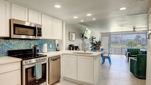 A modern kitchen with white cabinetry, a stainless steel stove, and a microwave. There is a dining area with a green chair and a view of the outdoors through large windows.