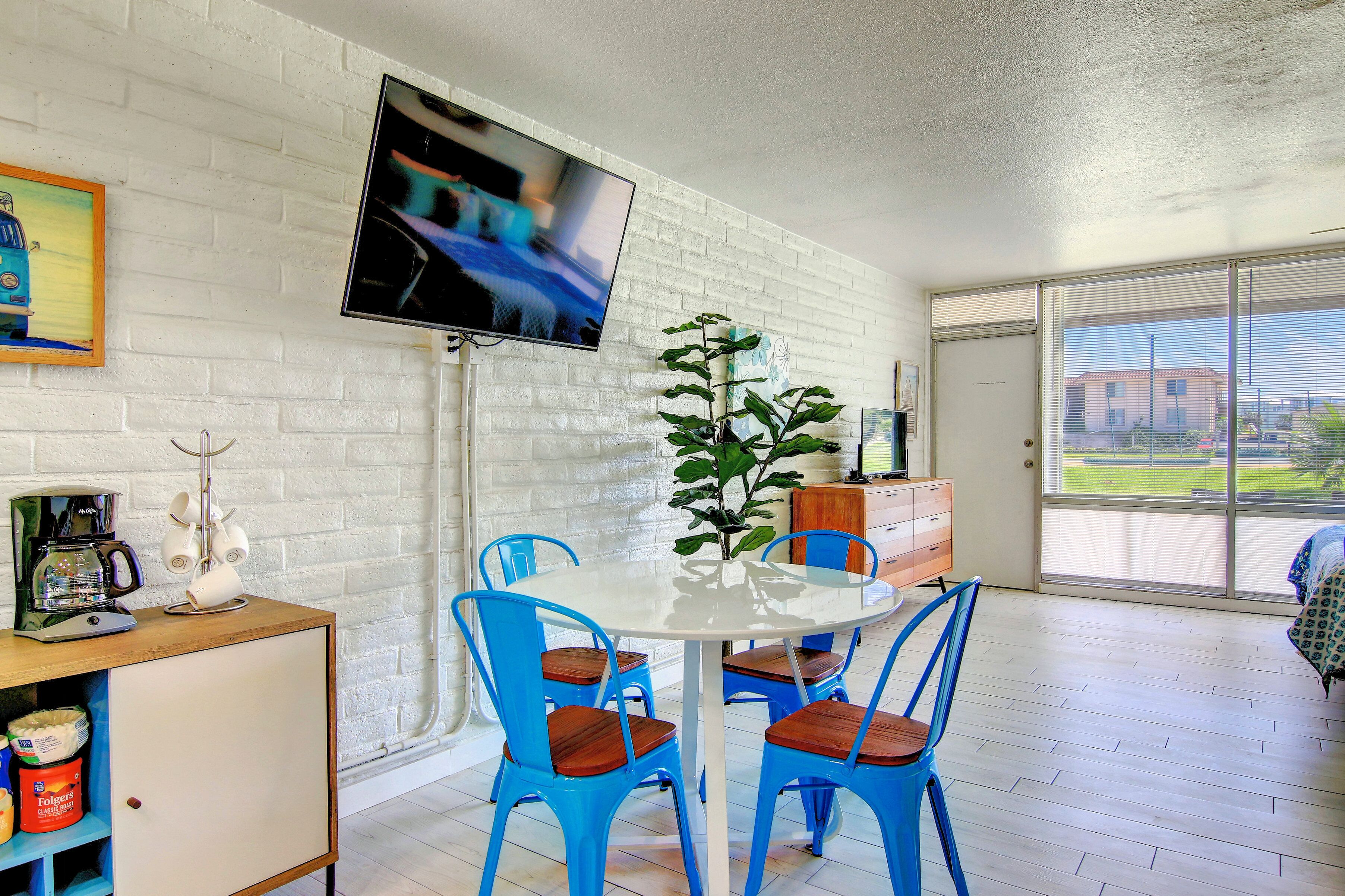 A room with a white brick wall, a flat-screen TV mounted above a white table surrounded by blue chairs, a potted plant, and a view through a large window.