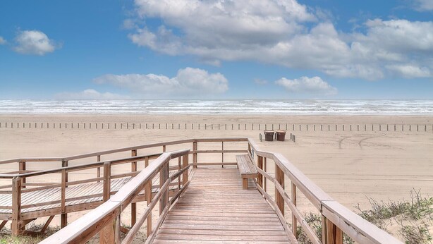 A wooden boardwalk leading to a sandy beach with a wooden railing, a bench, and two trash bins.