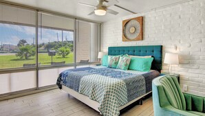 A bedroom with a large bed, a green armchair, a ceiling fan, and a view of a green lawn through floor-to-ceiling windows.