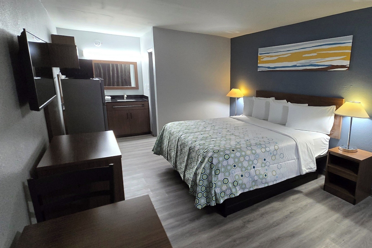 Habitación de hotel con cama, mesitas de noche, televisor montado en la pared y un cuadro sobre la cama.