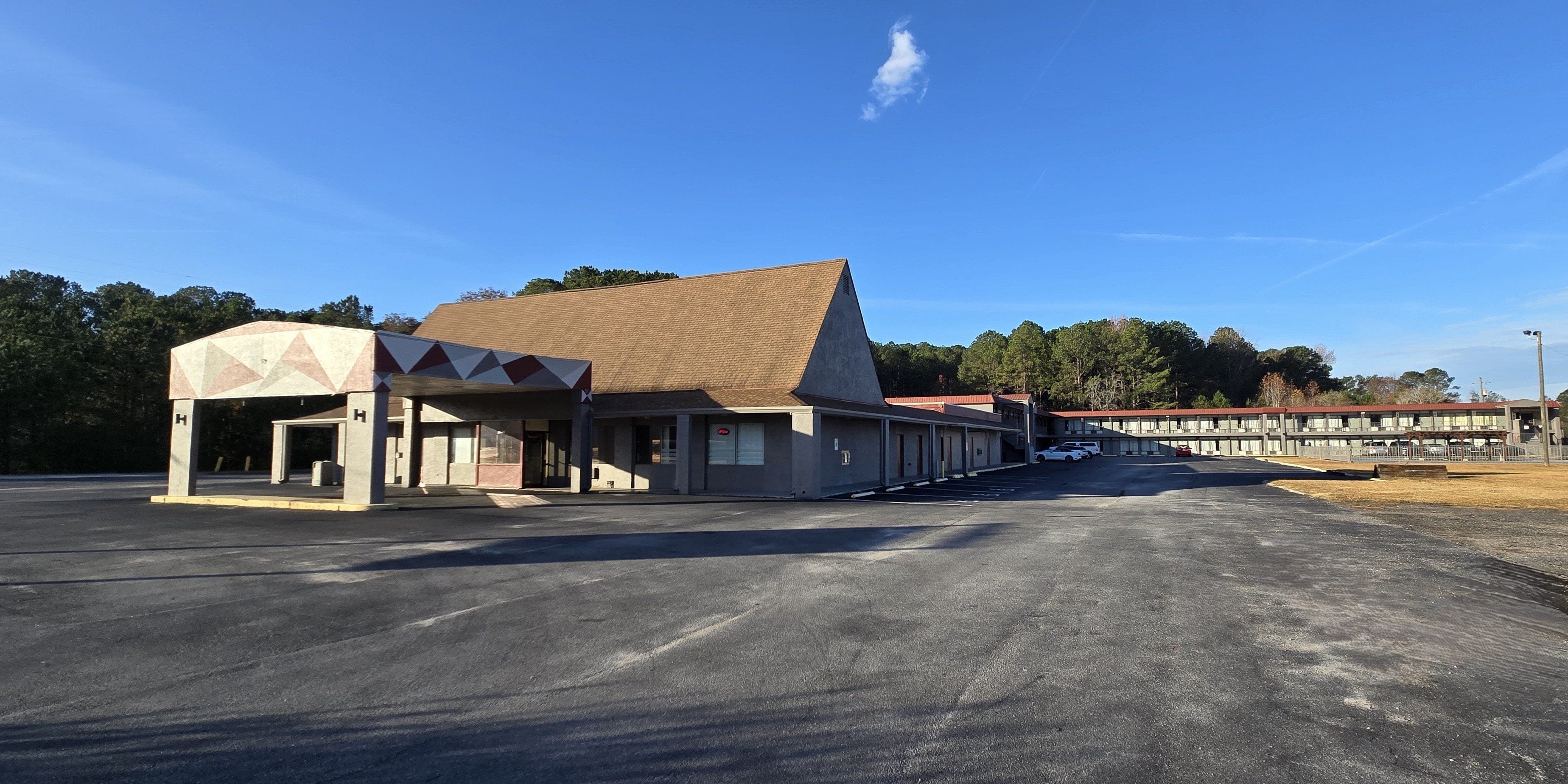A motel with a central building and attached wings, featuring a parking lot and a clear sky.