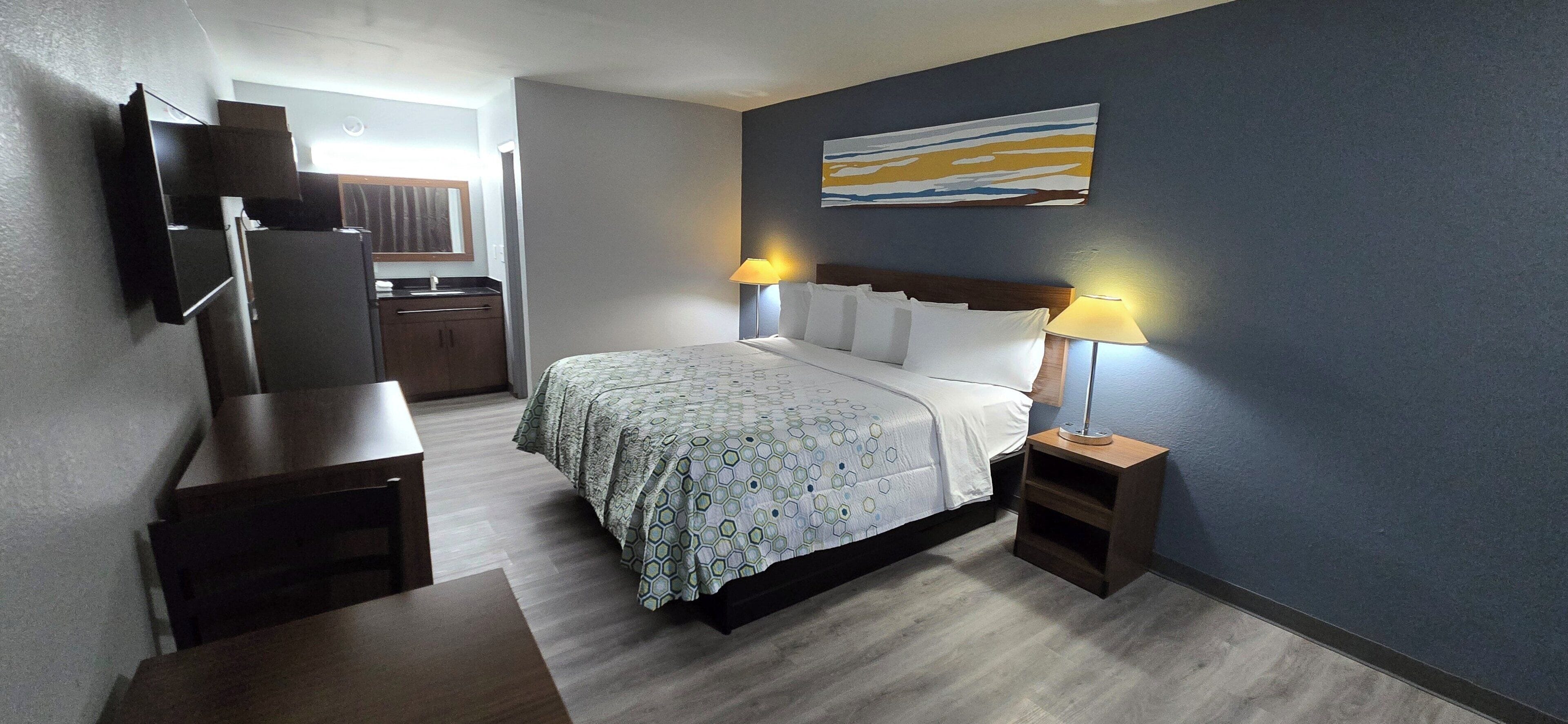 Habitación de hotel con cama, mesitas de noche, televisor montado en la pared y un cuadro sobre la cama.