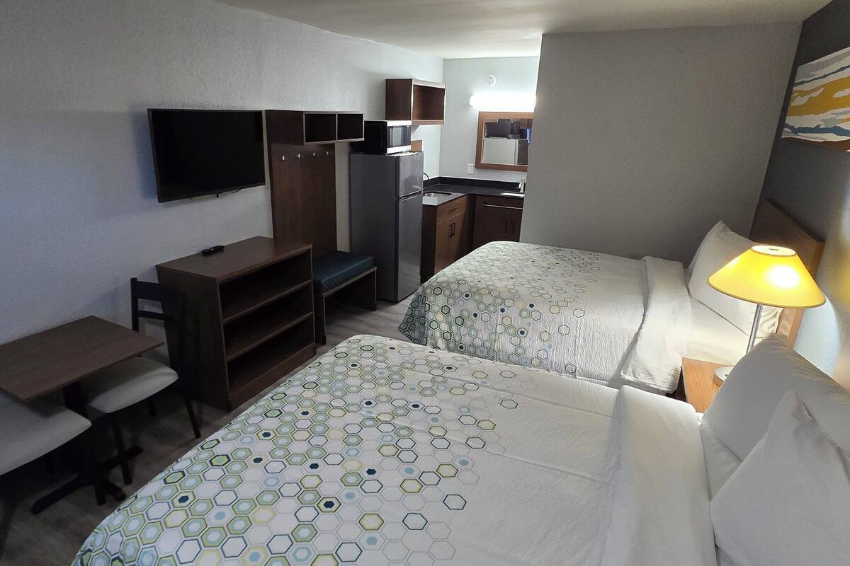 A hotel room with two beds, a desk, a chair, a TV, a kitchenette, and a painting on the wall.