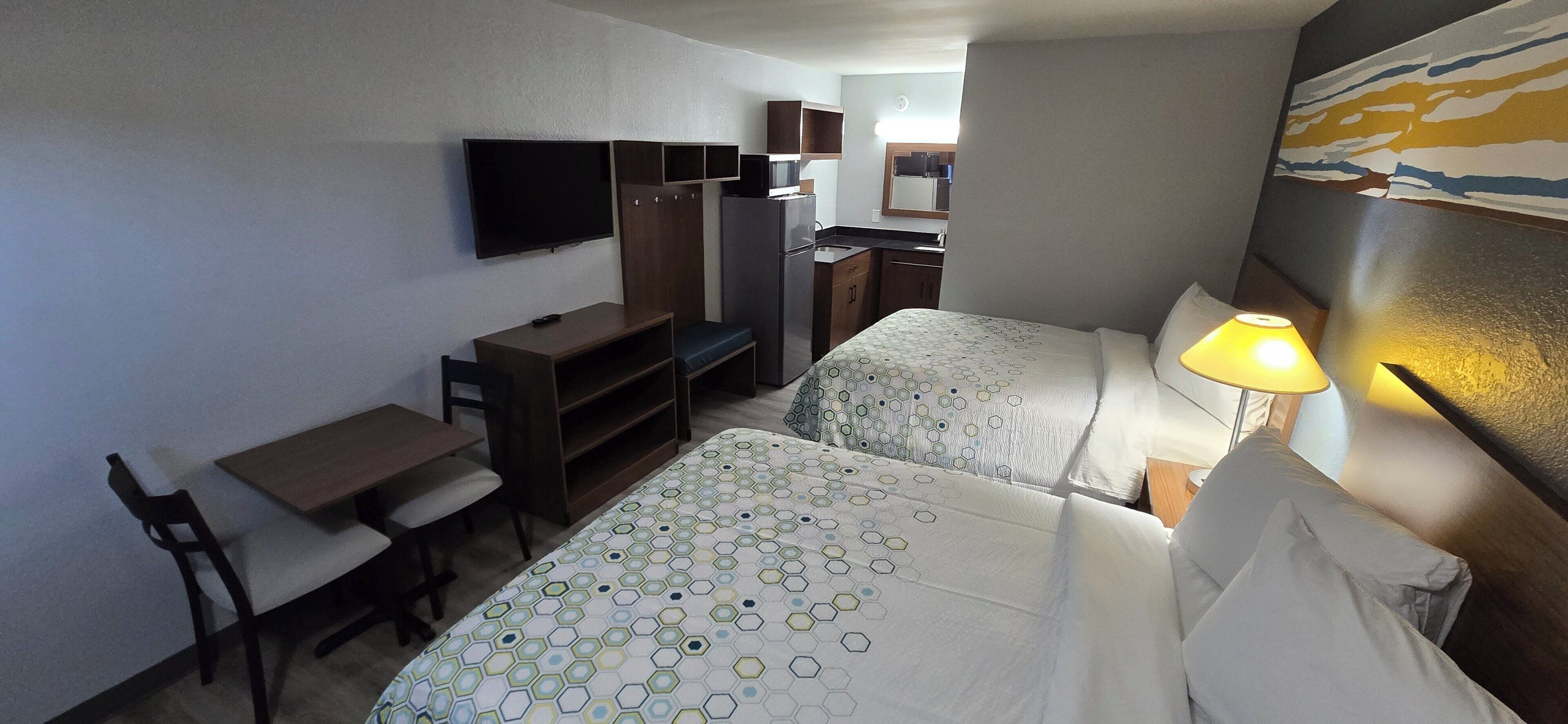 Habitación de hotel con dos camas, un escritorio, una silla, un televisor, kitchenette y un cuadro en la pared.