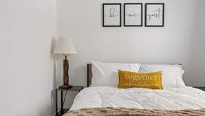 En pænt redt seng med hvidt sengetøj og et brunt tæppe, en sennepsgul pude med tekst, et natbord med en lampe, og tre billeder med inspirerende ord på væggen.