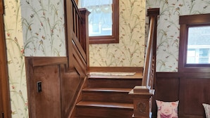Una escalera de madera con barandilla, flanqueada por un papel tapiz floral y un ventanal con cortina de encaje.