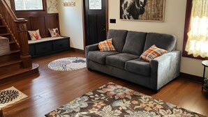 Una sala de estar con un sofá gris, una escalera de madera, una araña y un tapete grande con estampado floral.