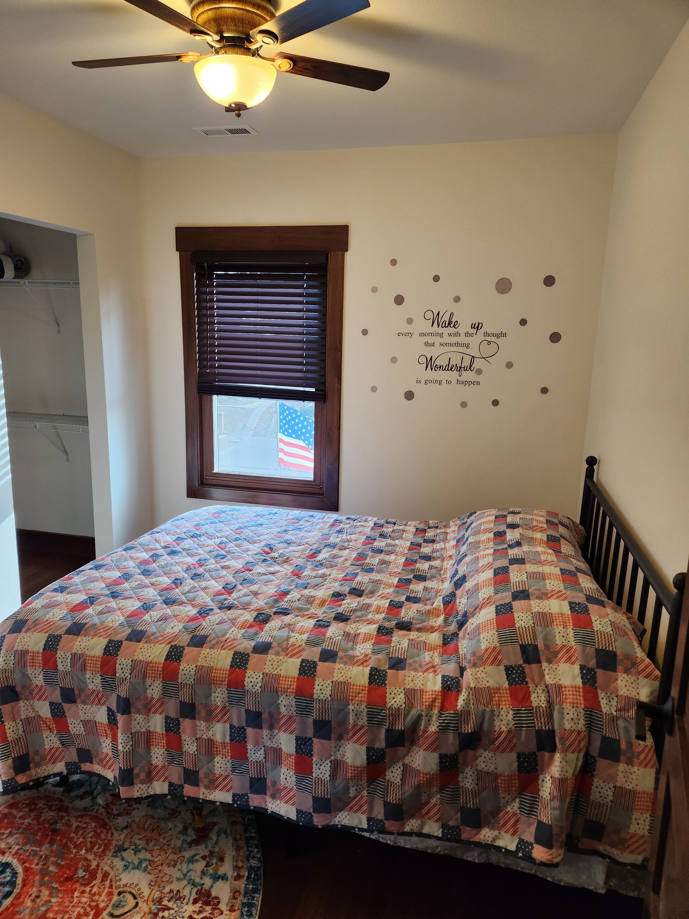 A bedroom with a bed, a ceiling fan, a window with blinds, and a motivational wall decal.