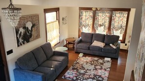 Un salón con un sofá azul, un sofá gris, una mesa de centro de madera y un tapete floral. Se observa una araña, un cuadro de un toro y una ventana con cortinas florales.