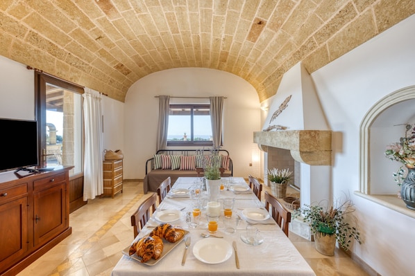 A dining area with a long table set for a meal, a television on a wooden cabinet, and a stone fireplace.