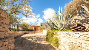 Un sentier de pierres mène à un bâtiment rustique en pierre, doté d’un toit en tuiles, et est entouré de verdure et d’un agave.