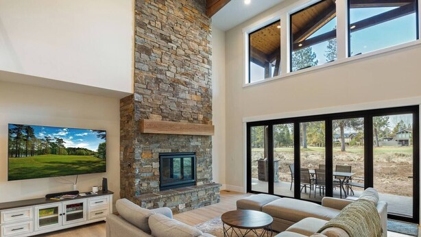 A spacious living room with a stone fireplace, a large flat-screen TV, a sectional sofa, and a view of the outdoors through large sliding glass doors. - Sleeps 16+ | 7 bed 7.5 bath | EV | Hot tub | SHARC (Sunriver)