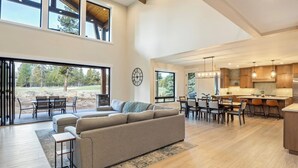 A spacious living area with a large sofa, dining table, and chairs, a kitchen with wooden cabinets, and large windows offering a view of the outdoors. - Sleeps 16+ | 7 bed 7.5 bath | EV | Hot tub | SHARC (Sunriver)