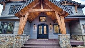 A two-story house with a stone foundation, a dark front door, and a wooden porch with a triangular roof structure. - Sleeps 16+ | 7 bed 7.5 bath | EV | Hot tub | SHARC (Sunriver)