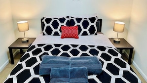 Un lit orné d’un motif géométrique noir et blanc, deux tables de chevet avec des lampes, un coussin rouge et un élément décoratif mural au-dessus du lit.