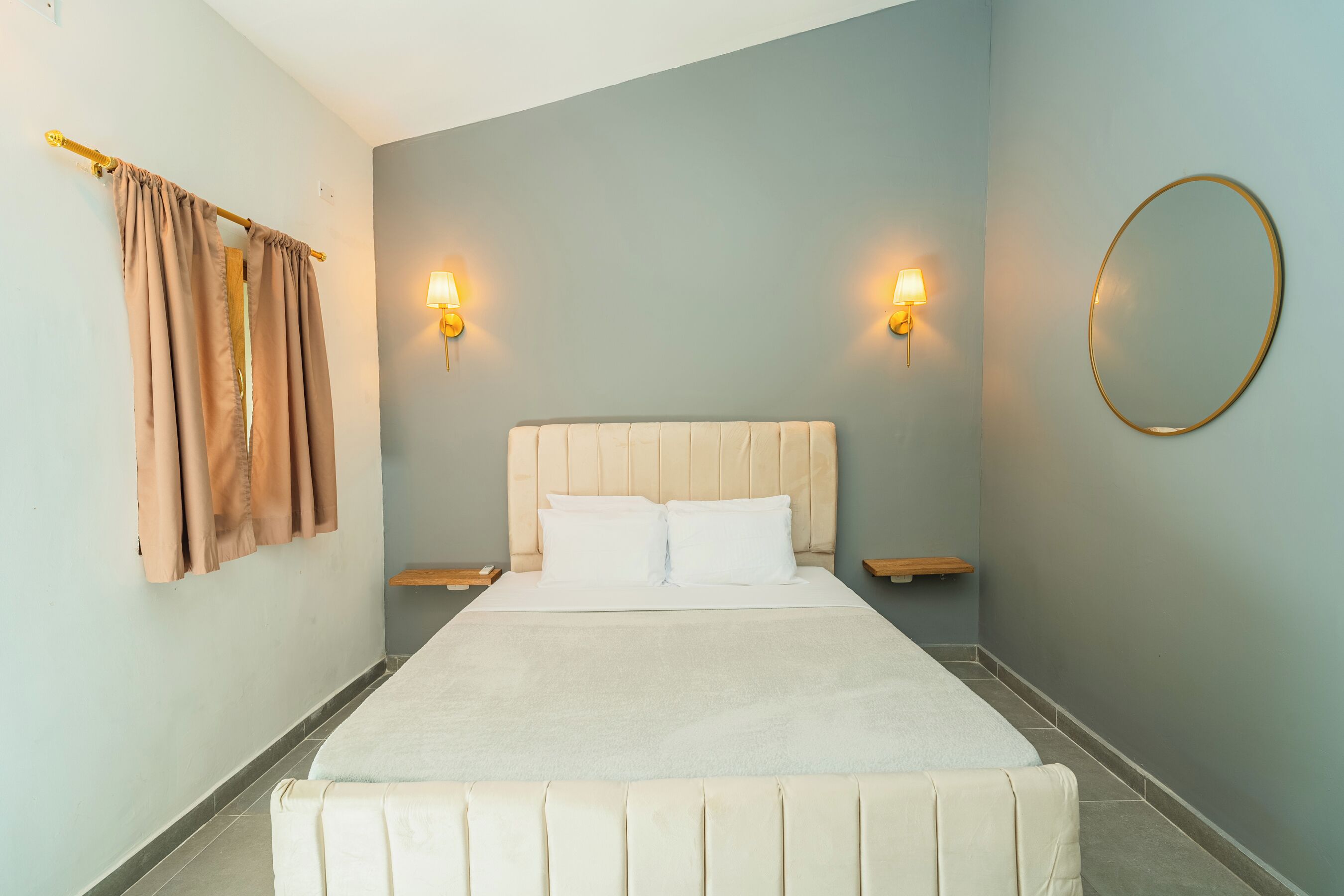 A neatly made bed with white linens, two bedside tables with lamps, a round mirror on the wall, and a curtain on the left.