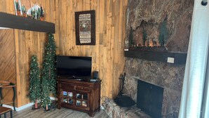 A cozy living room with a wooden wall, a flat-screen TV, a Christmas tree, a fireplace, and a stone accent wall. - Cozy 3-bedroom condo with fire place and WiFi in Fraser, CO (Fraser)