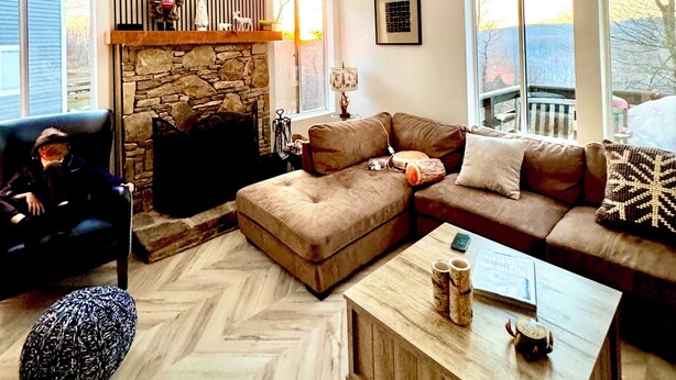 A spacious living room with a stone fireplace, a large window, and a high ceiling with exposed beams.
