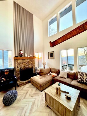 Un salón amplio con chimenea de piedra, un ventanal grande y un techo alto con vigas a la vista.