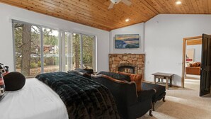 Une chambre confortable avec une cheminée en pierre, un lit recouvert d’une couverture sombre, un fauteuil noir et une vue dégagée sur l’extérieur grâce à de grandes fenêtres.