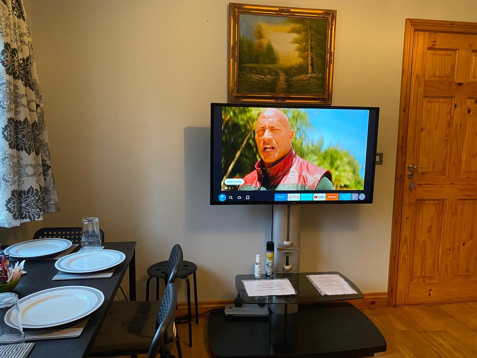A room with a television mounted on the wall, a dining table set with plates and cutlery, a chair, and a wooden door.