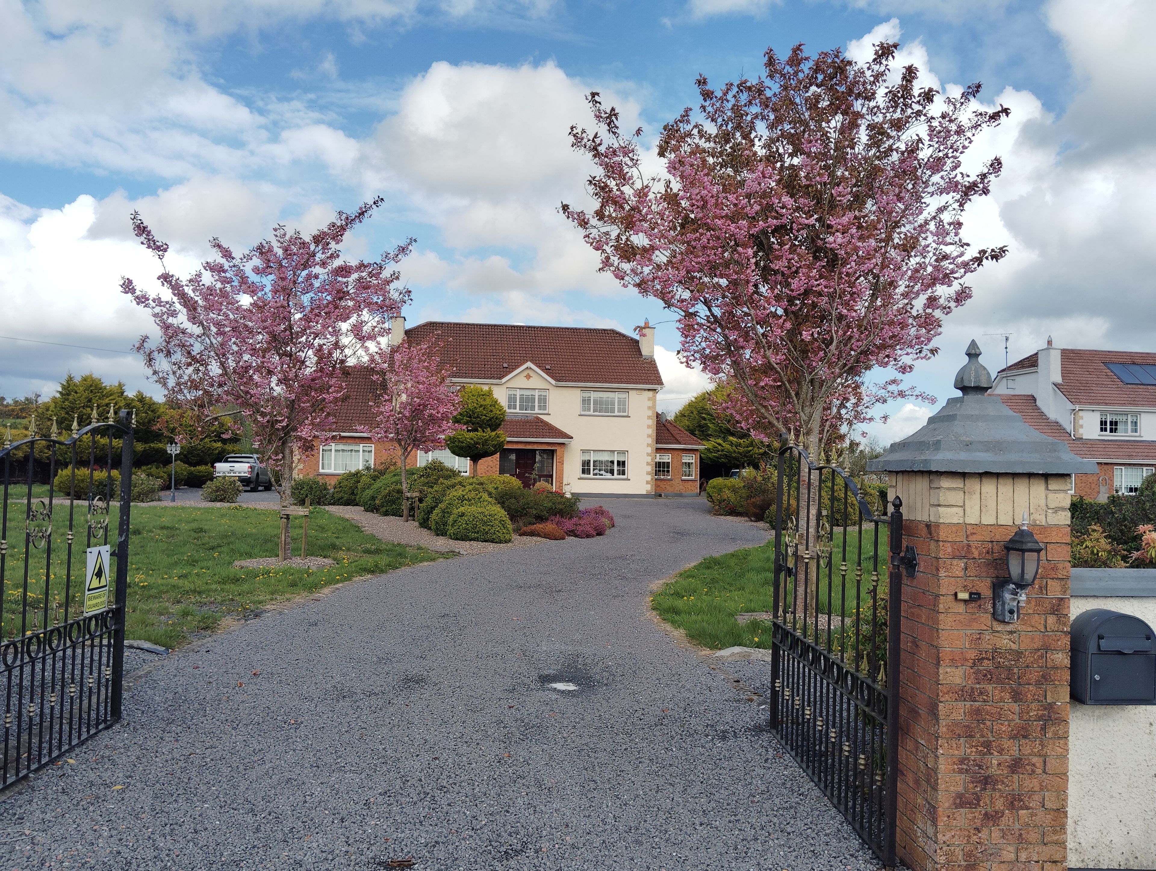A residential driveway leading to a house with a gravel path, surrounded by flowering trees and a gated entrance.