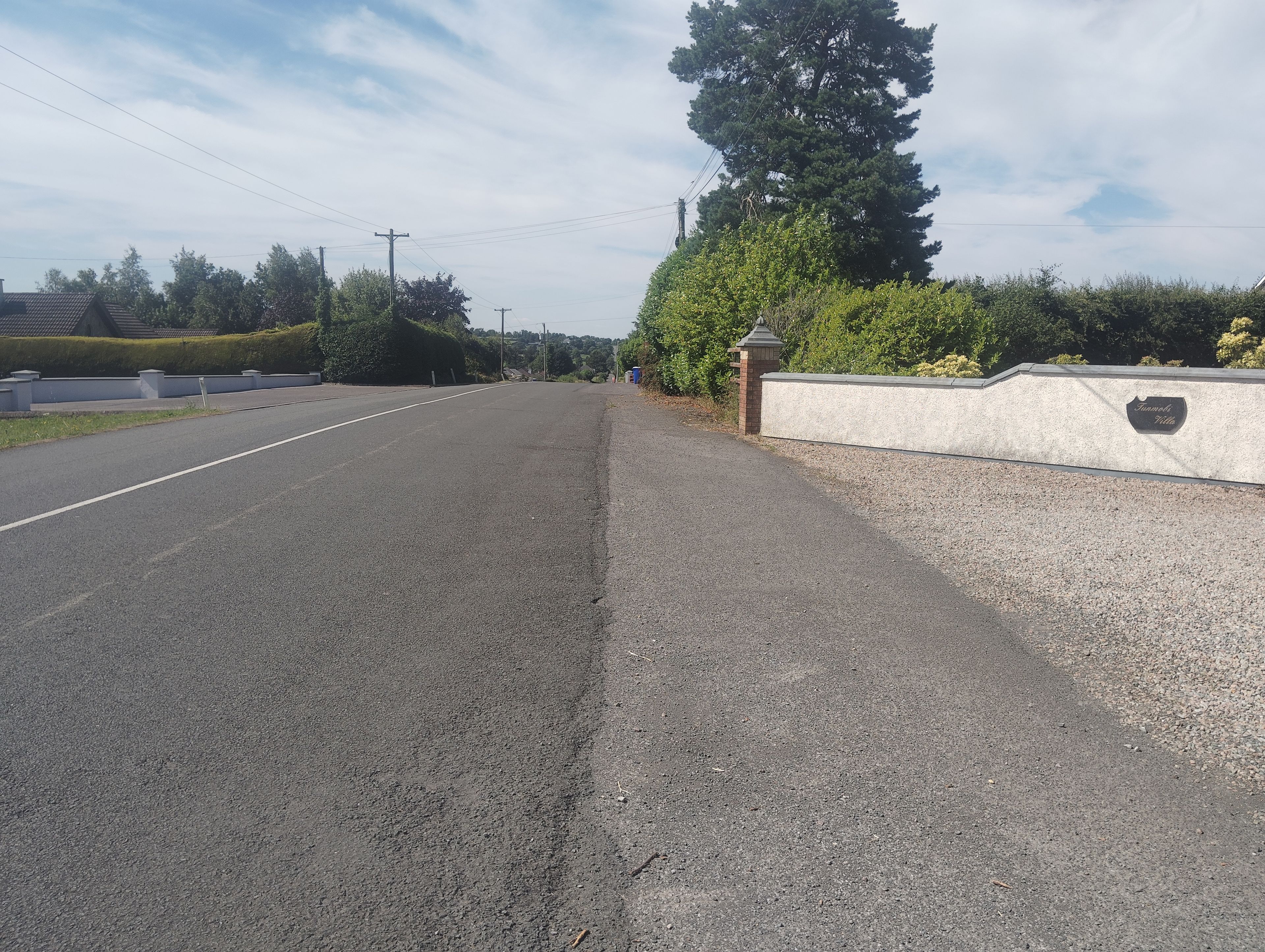 Une route tranquille, bordée d’une bande de gravier, d’un mur de pierres et d’un panneau indiquant « Sunnyside Villas ».