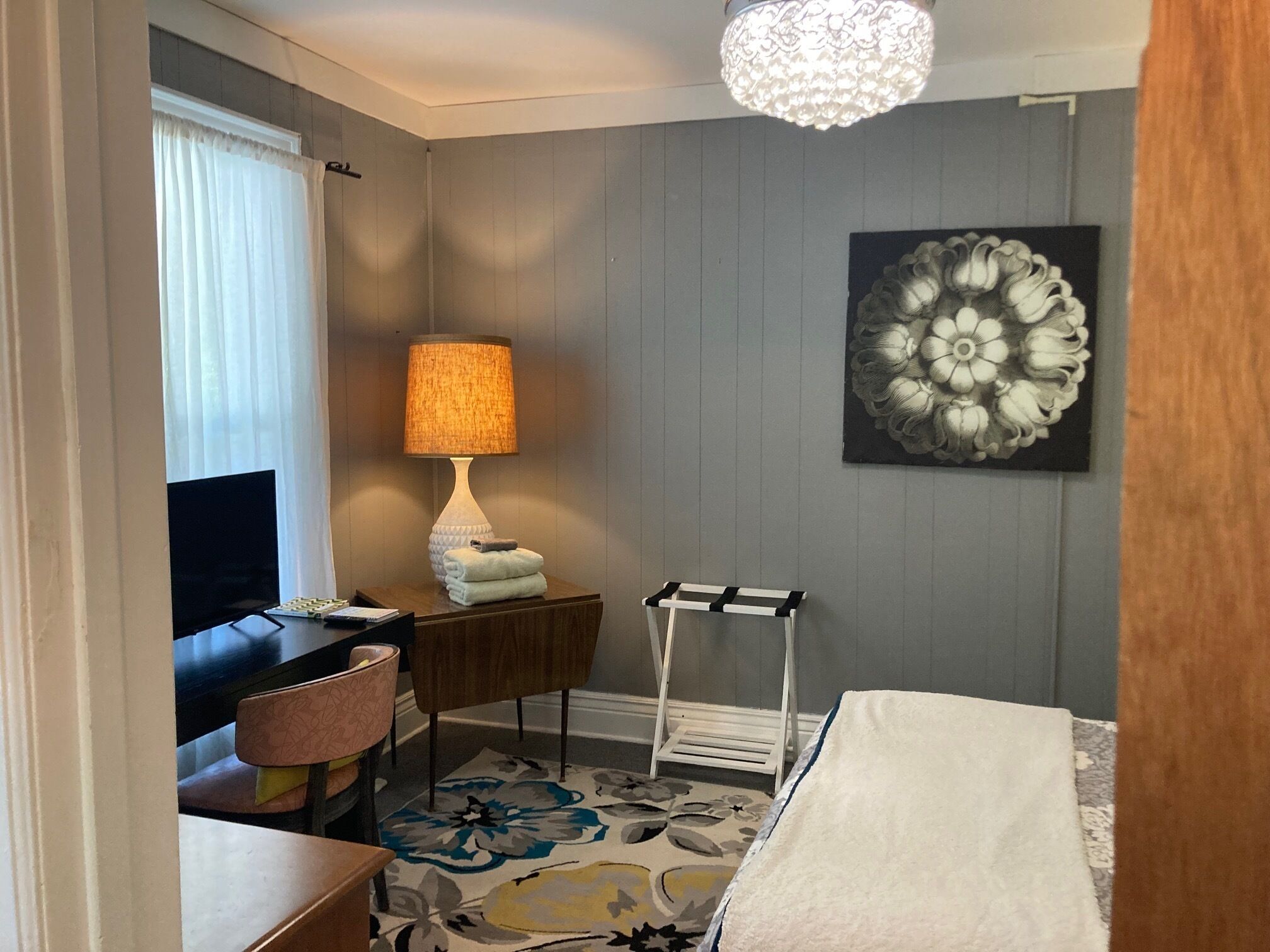 A hotel room with a bed, a desk with a lamp, a chair, a small table, a decorative wall piece, and a window with curtains.