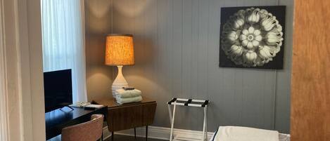 A hotel room with a bed, a desk with a lamp, a chair, a small table, a decorative wall piece, and a window with curtains.