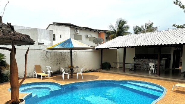 A swimming pool with a tiled deck, surrounded by white chairs and a covered patio area. - Hotel Lunna (Porto Nacional)