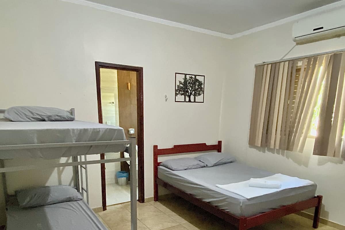 Una habitació de dormitori amb literes, un llit individual, una porta, un aire condicionat fixat a la paret i una finestra amb cortines.