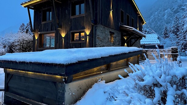 Una cabaña de madera con techo nevado y una terraza cubierta de nieve al frente.