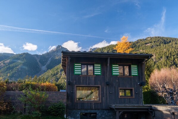 A two-story wooden house with green shutters, set against a mountainous backdrop.