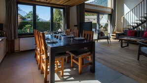 Un comedor con una mesa de madera dispuesta para seis personas, rodeada de sillas de madera. Se ve un sofá y un televisor al fondo.