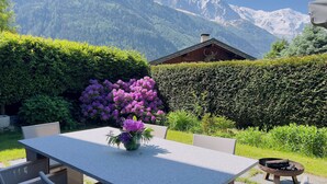 Una mesa con sillas, un florero y una chimenea en un jardín con montañas al fondo.