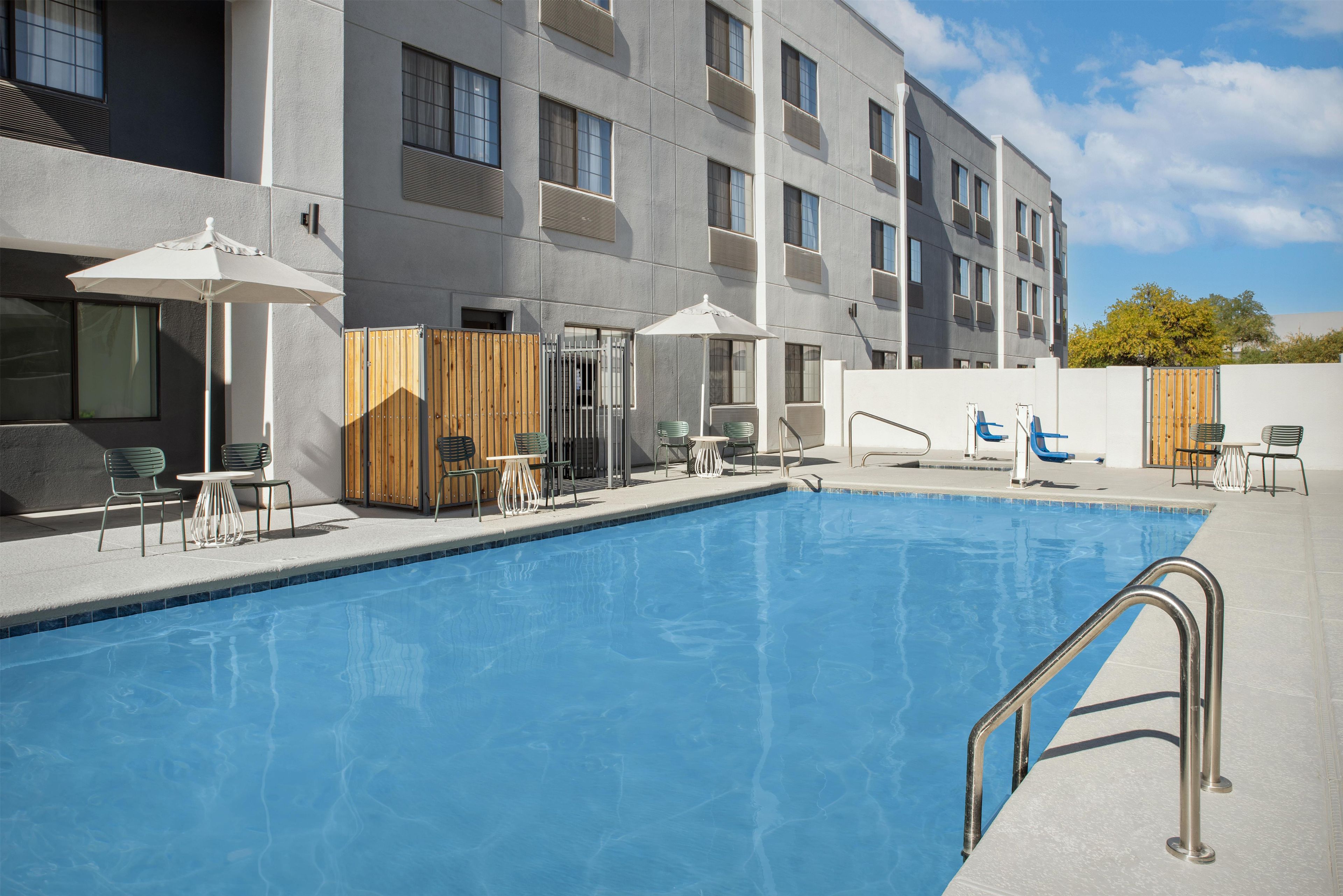 A swimming pool with a metal ladder, surrounded by lounge chairs and umbrellas, with a building in the background.