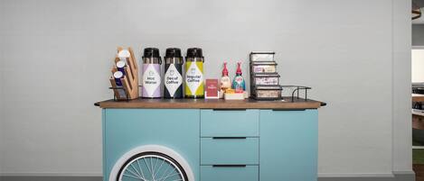 A teal cart with beverage dispensers labeled "Hot Water," "Decaf Coffee," and "Regular Coffee," along with a small display of Folgers coffee and other items.
