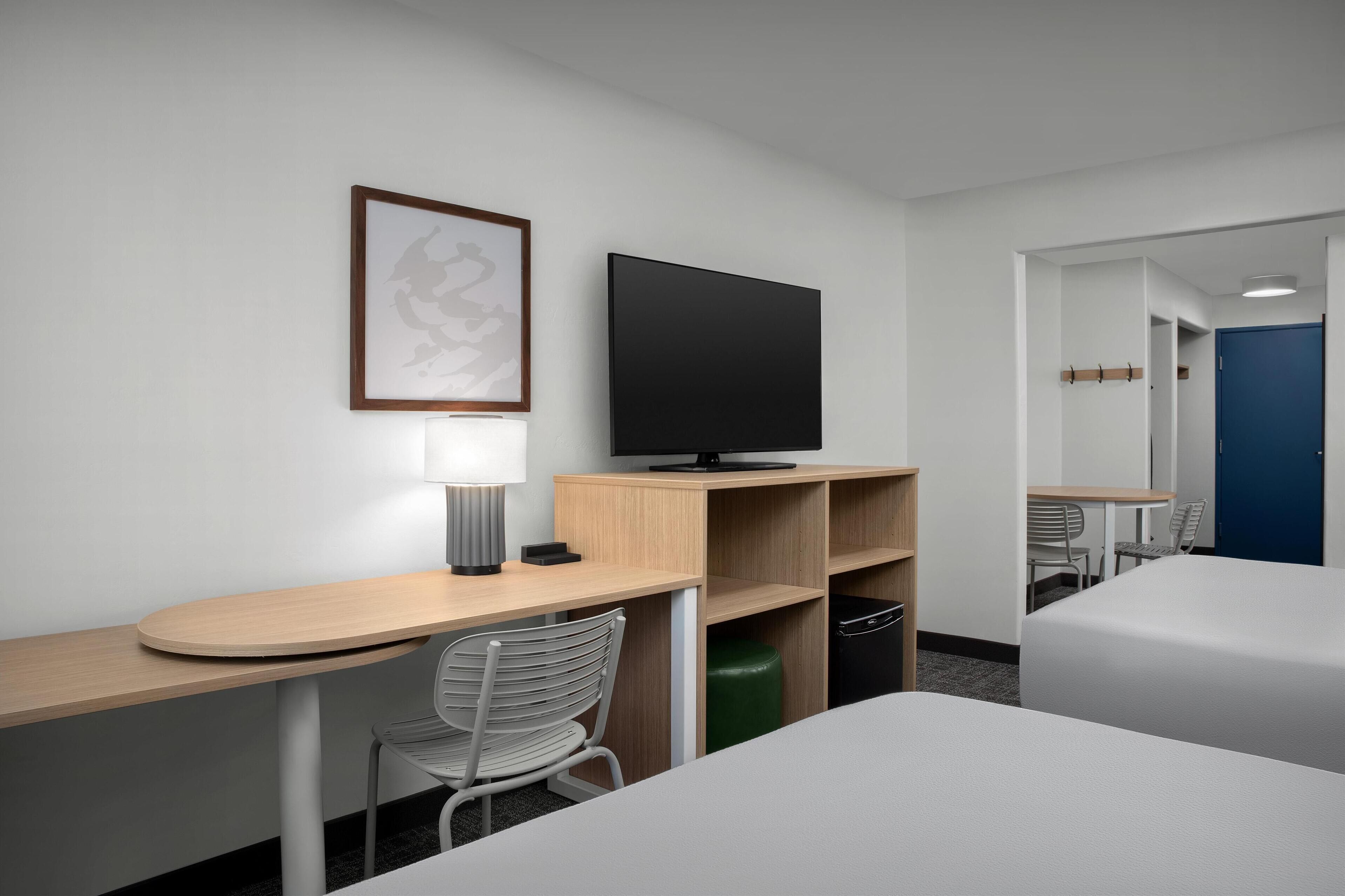 Ein Hotelzimmer mit einem Bett, einem Schreibtisch mit Stuhl, einem Fernseher, einer Lampe und einem gerahmten Bild an der Wand.