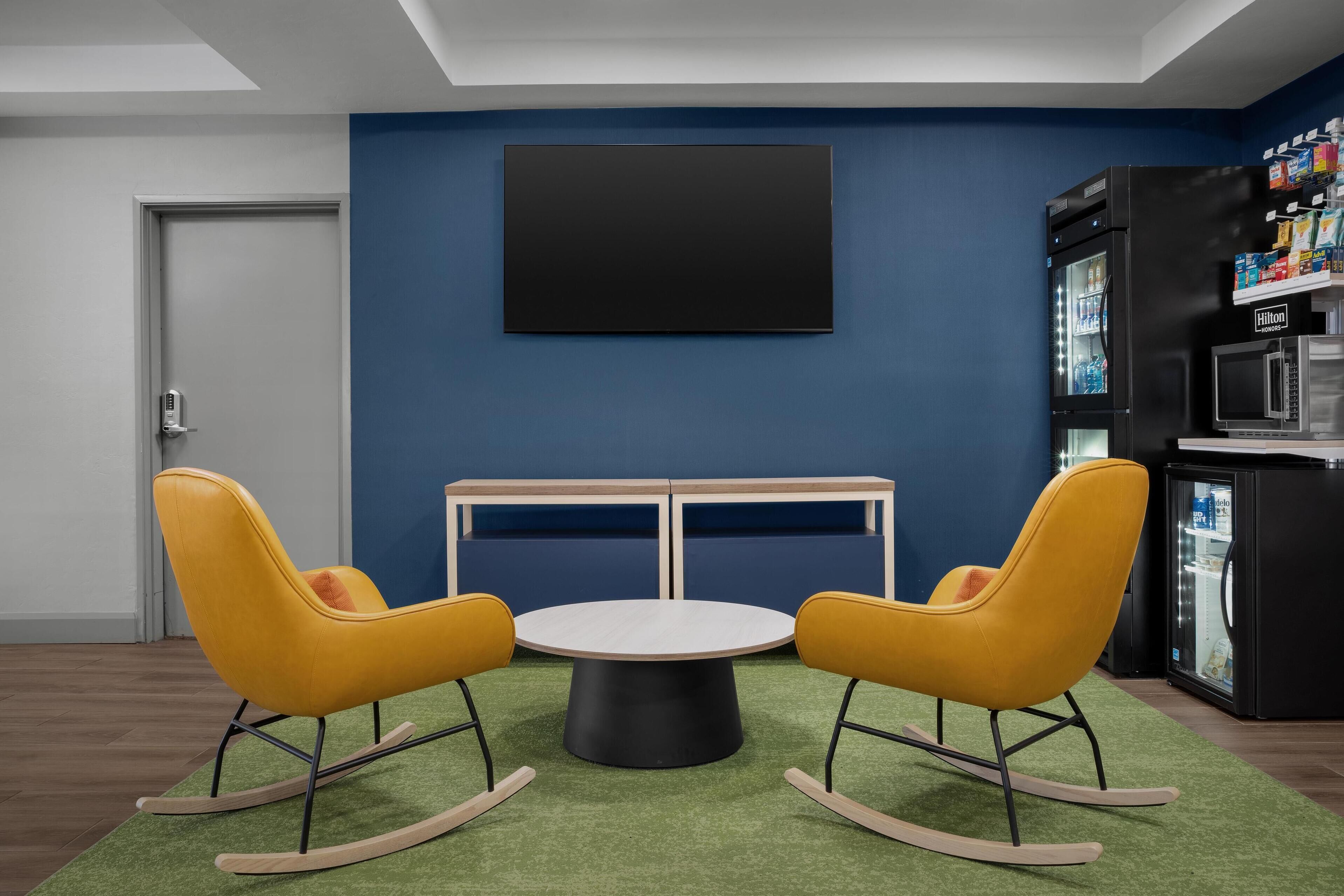 A modern room with a blue wall, a black TV mounted above a wooden table with two yellow rocking chairs, and a minibar with a refrigerator and microwave.