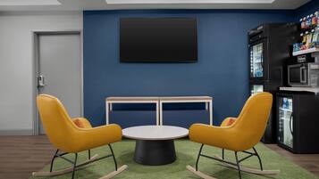 A modern room with a blue wall, a black TV mounted above a wooden table with two yellow rocking chairs, and a minibar with a refrigerator and microwave.